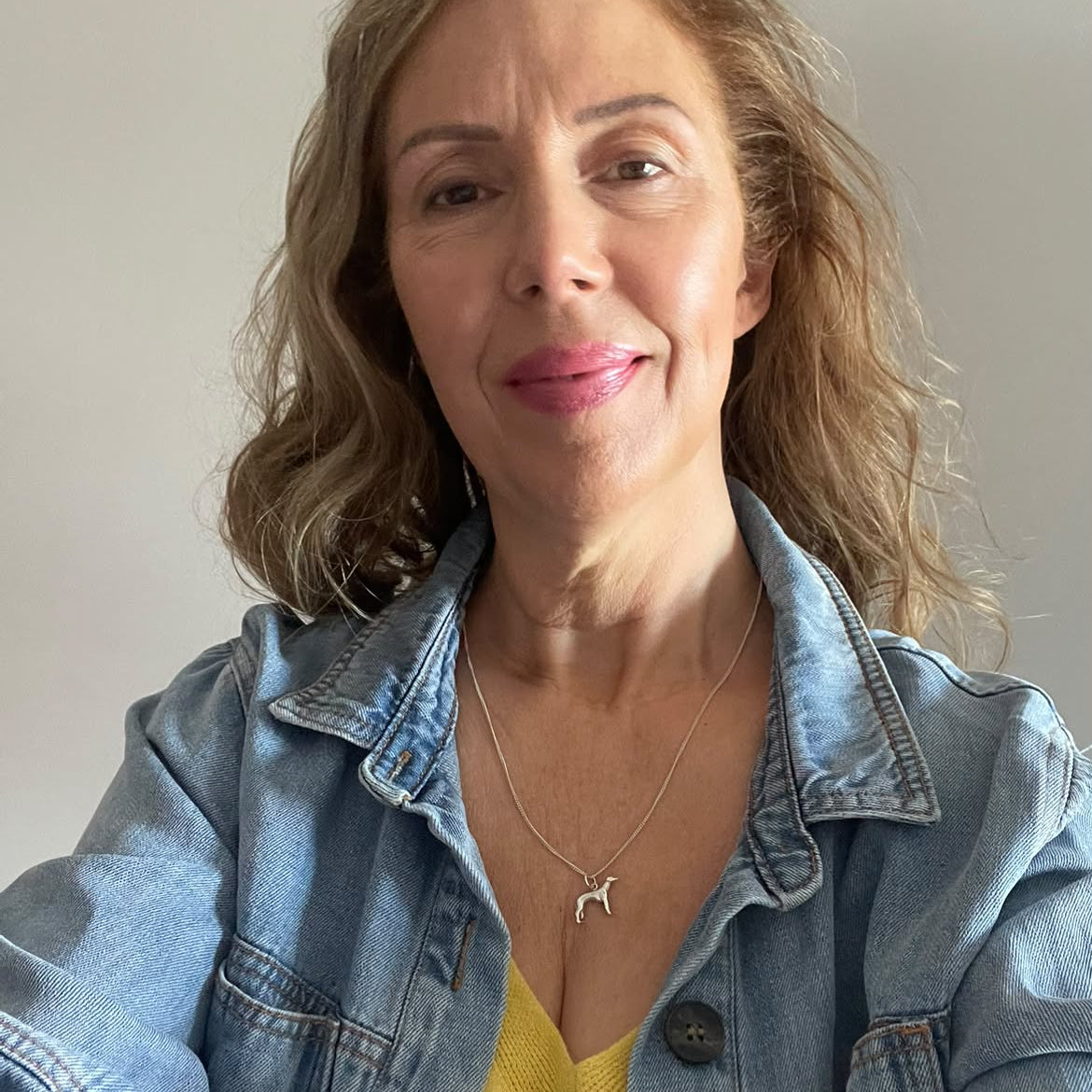 A woman with blonde wavy hair wearing a light blue denim jacket over a yellow top, accessorized with a silver greyhound necklace, photographed against a plain light gray wall.