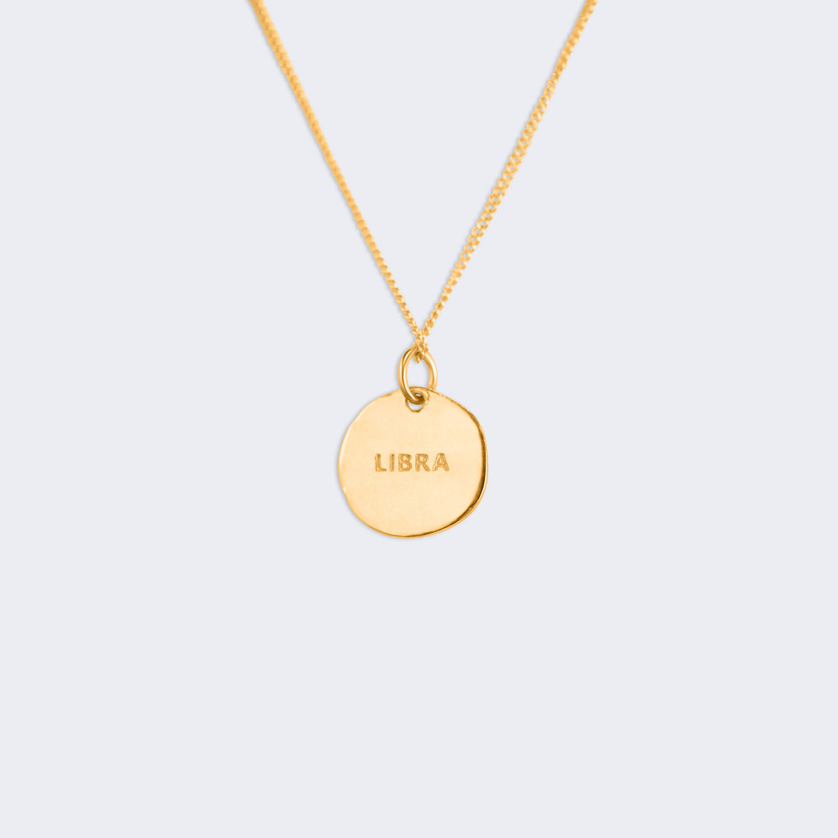 A gold chain necklace with a round gold disc pendant engraved with the word 'LIBRA', shown against a light gray background.