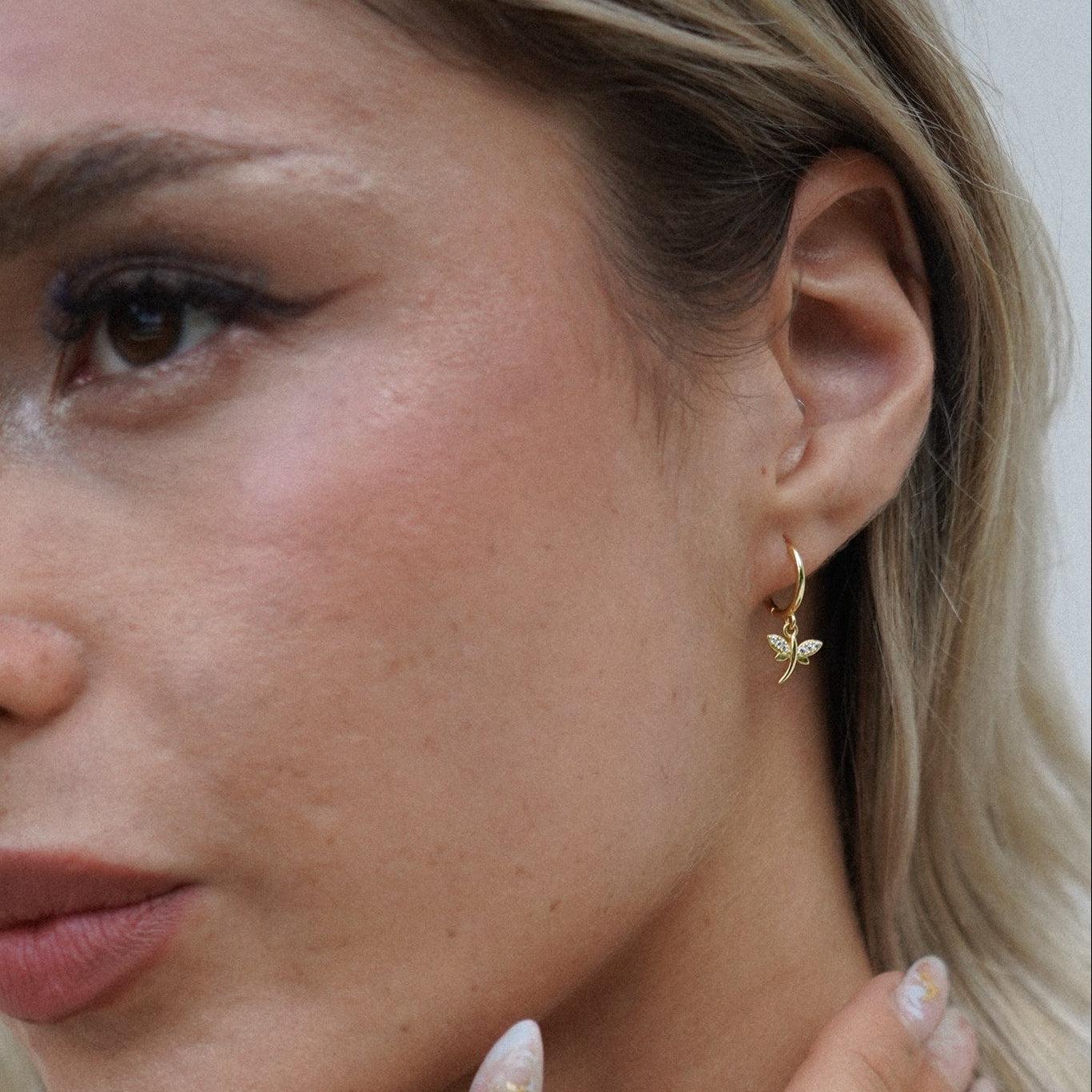 Close-up of a woman wearing small gold huggie earrings with a dragonfly charm on them