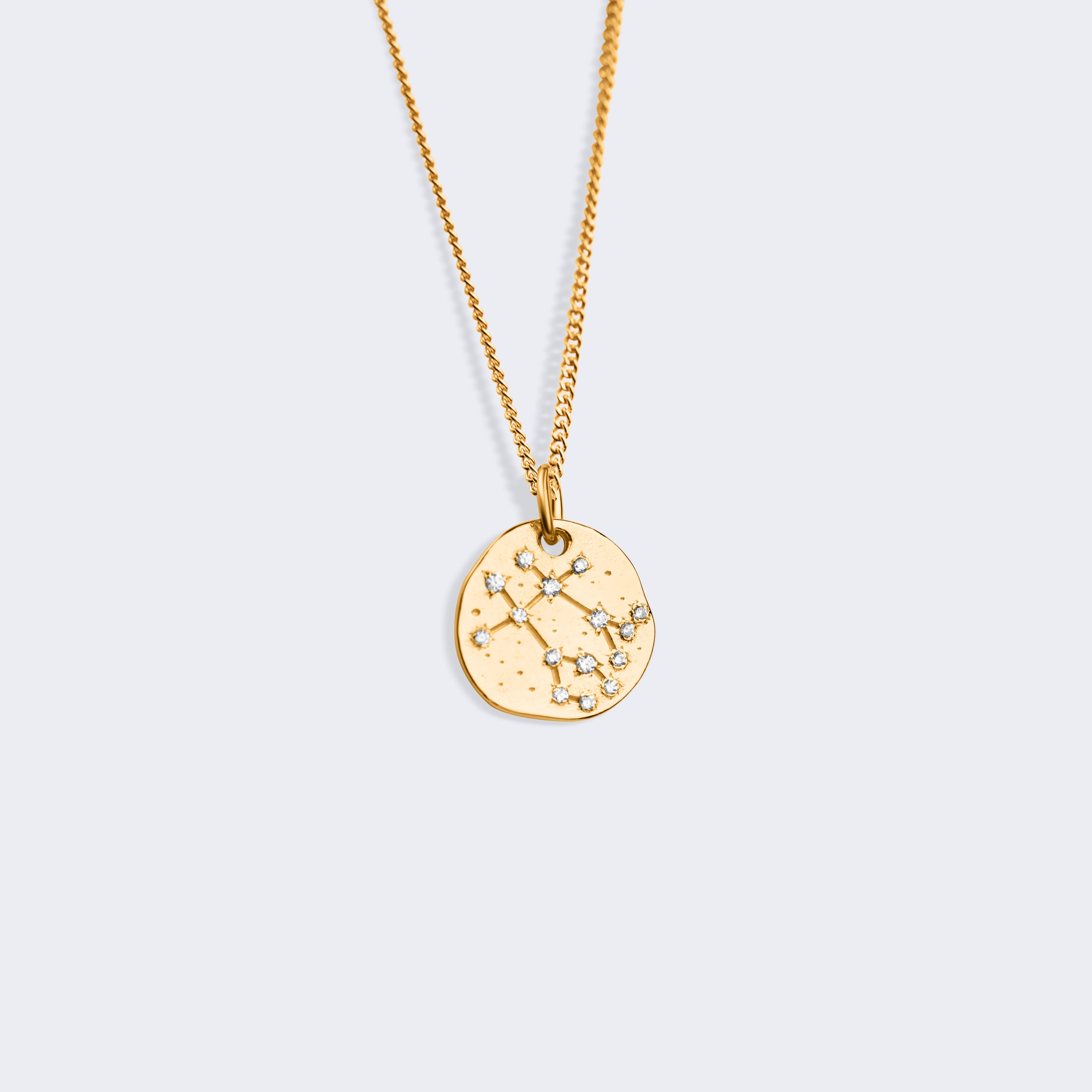 A gold chain necklace with a small, irregularly shaped gold disc pendant featuring an engraved celestial or zodiac design, star constellations of the gemini zodiac