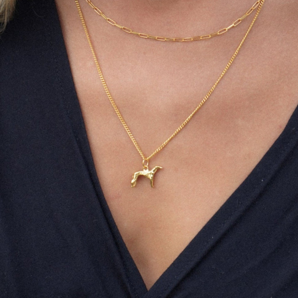 Close-up of a woman wearing a gold necklace with a small greyhound dog pendant against a neutral background