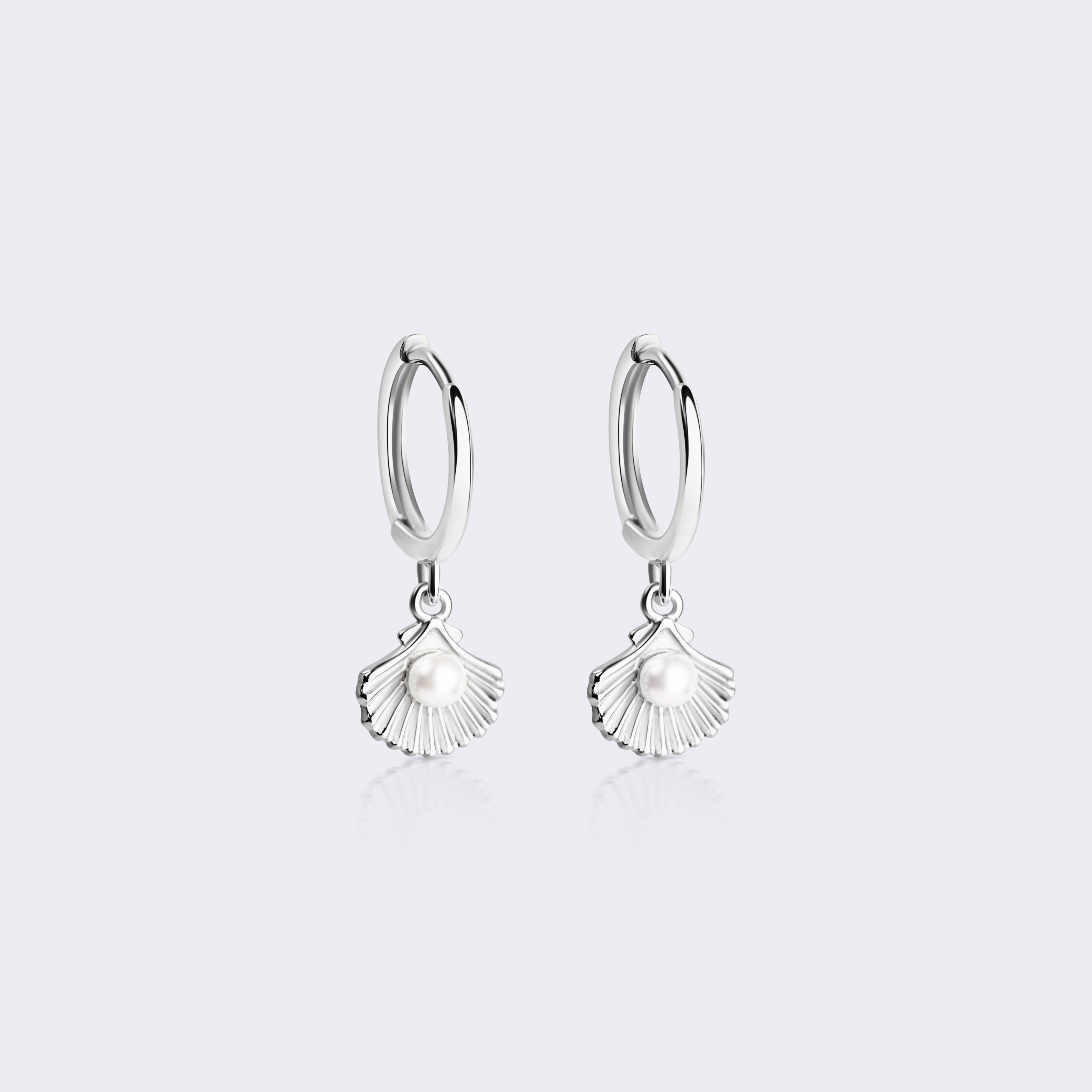 A pair of silver huggie hoop earrings with dangling scallop shell charms, shown on a light gray background.