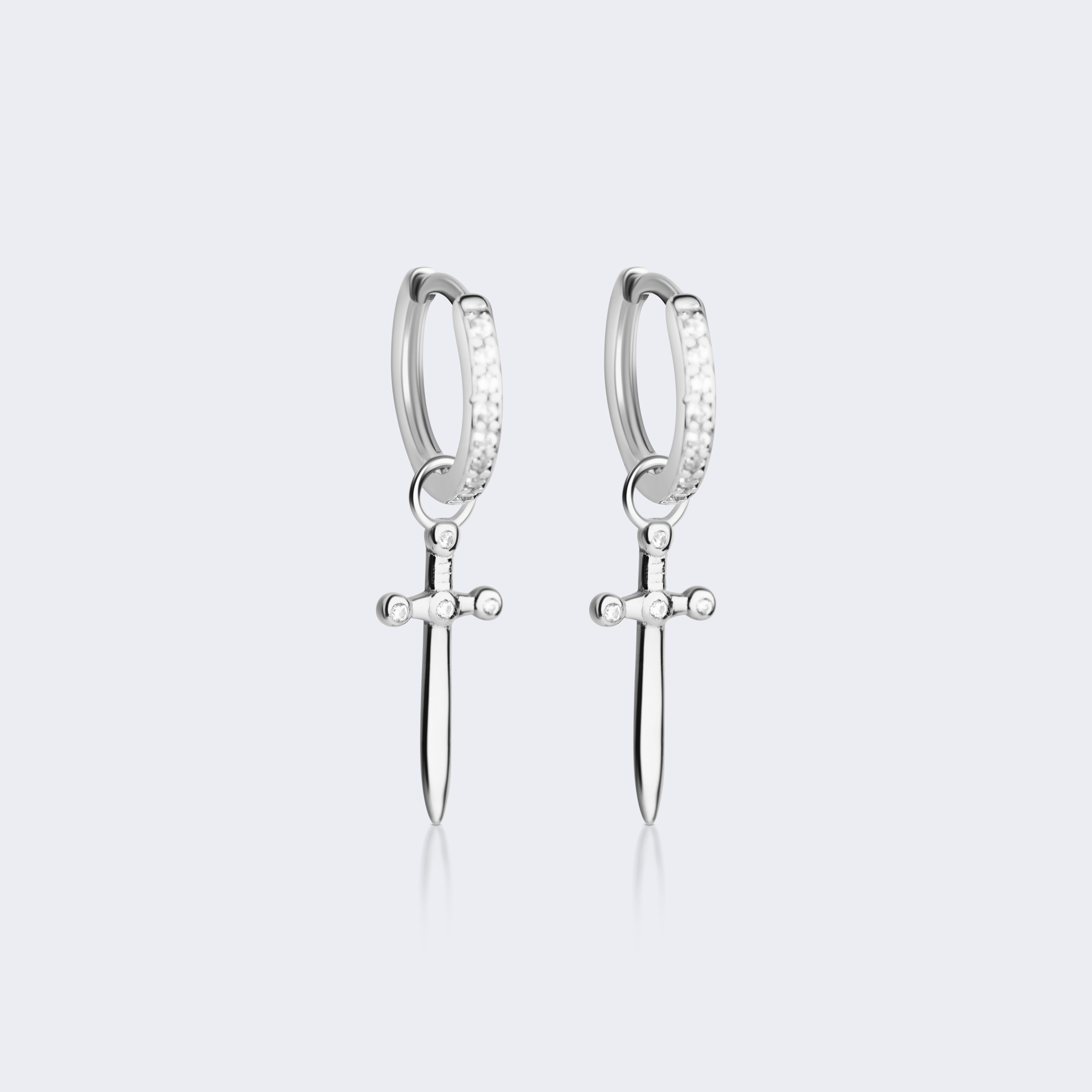 A pair of silver huggie hoop earrings with a dangling sword charm, set against a light gray background.