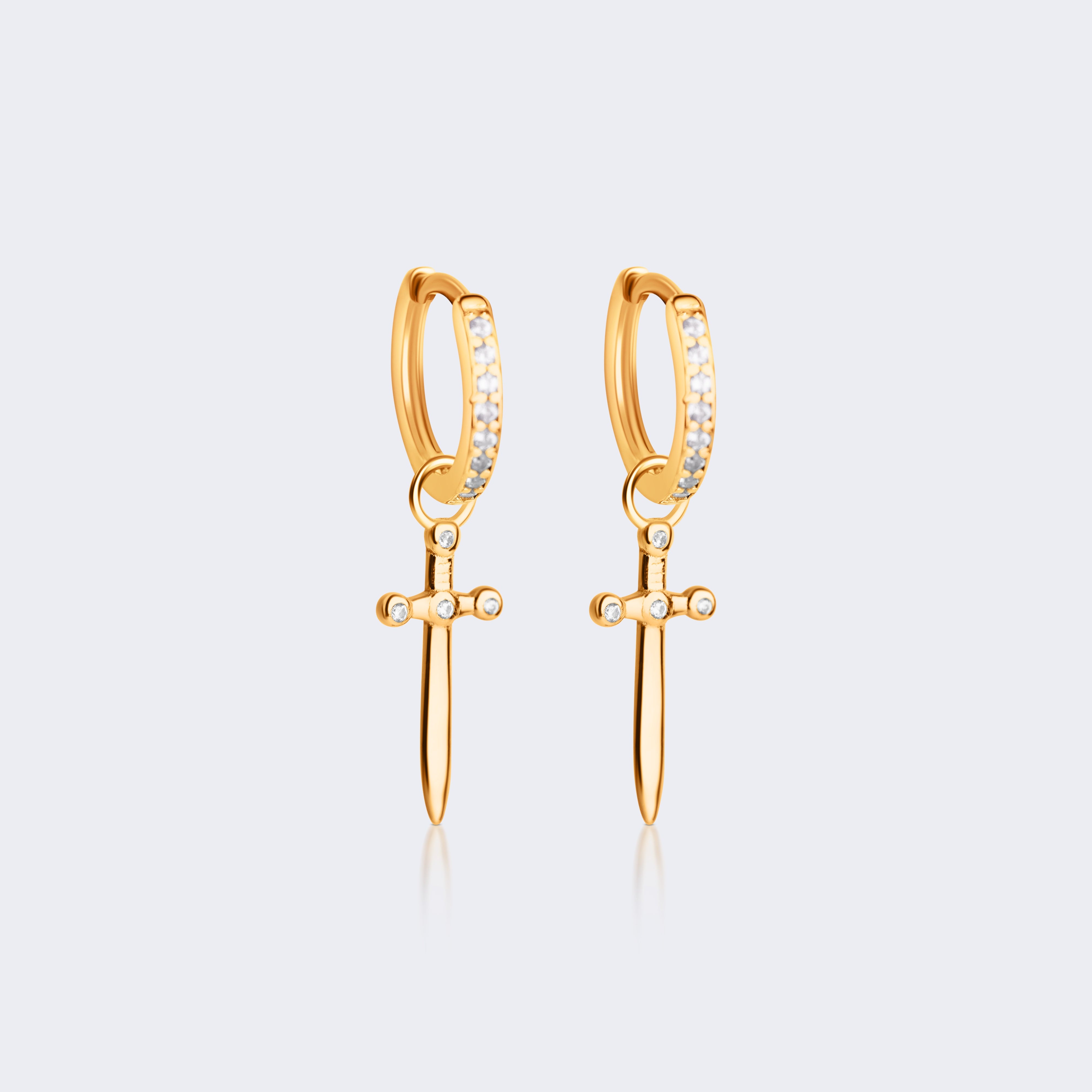 A pair of gold huggie hoop earrings with a dangling sword charm and a row of small clear stones on the front, shown on a light gray background.