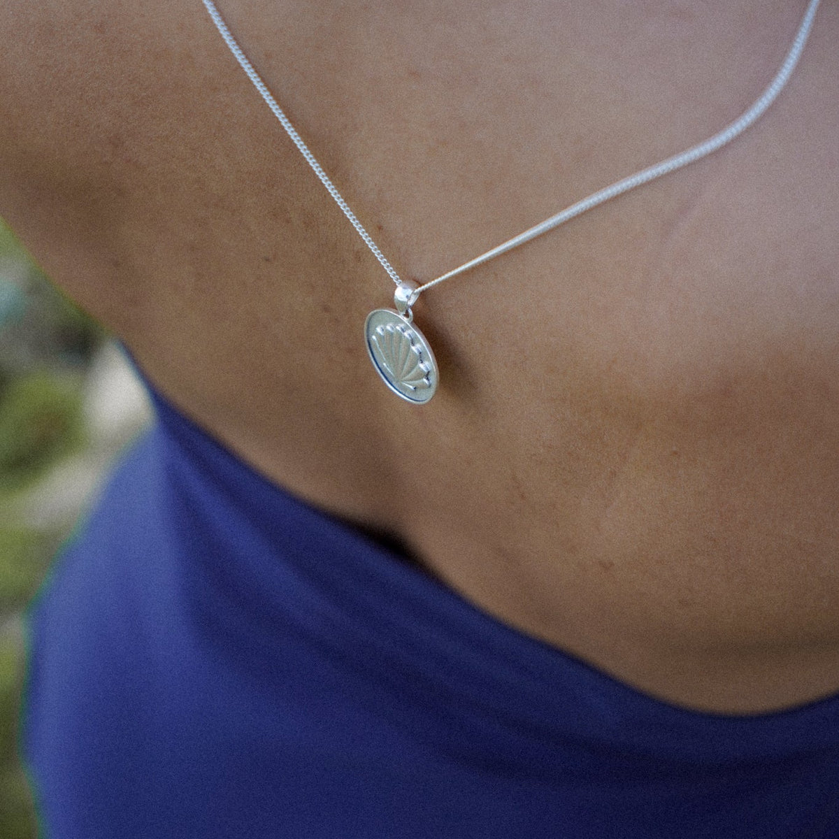 A close-up lifestyle shot of a person's bare back wearing a delicate silver chain necklace with a small oval pendant, paired with a blue halter top.