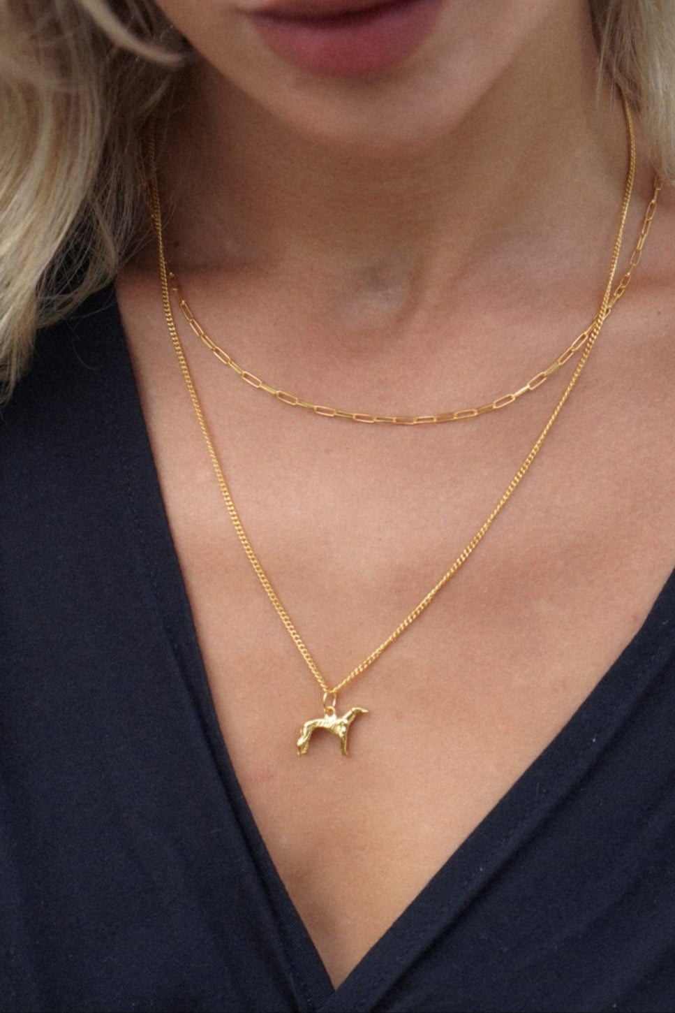 Close-up of a woman wearing a gold necklace with a small greyhound dog pendant against a neutral background